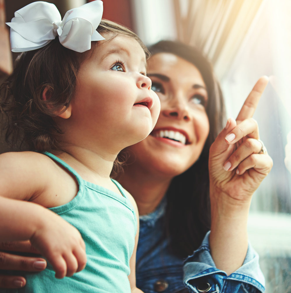 This is an image of a happy mom and toddler girl smiling and looking out a window.